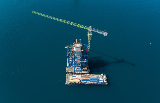 Aerial view of a towering crane rises from a barge, casting a stark silhouette against the tranquil, deep blue sea, Komarna, Dubrovnik-Neretva County, Croatia.