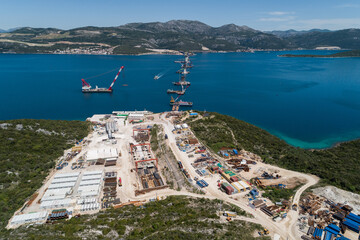 Aerial view of construction site sprawling with heavy machinery and half-finished bridge piers reaching into the azure sea, Komarna, Dubrovnik-Neretva County, Croatia.