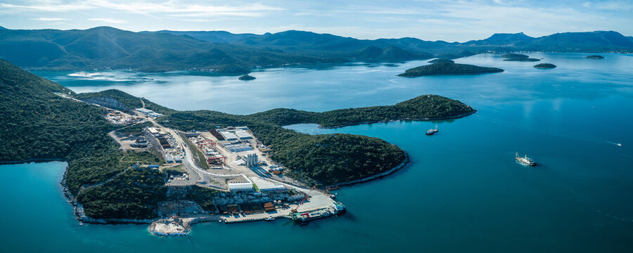 Aerial view of a coastal industrial area nestled between the deep blue sea and lush green hills, Komarna, Dubrovnik-Neretva County, Croatia.