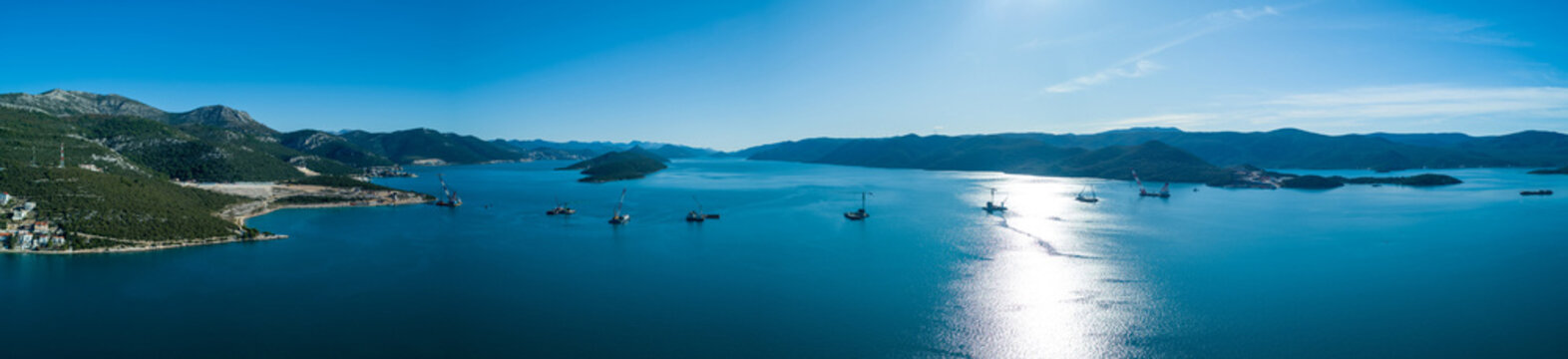Aerial view of cranes stand silhouetted against the shimmering turquoise sea, reflecting the bright sky in Komarna, Dubrovnik-Neretva County, Croatia.