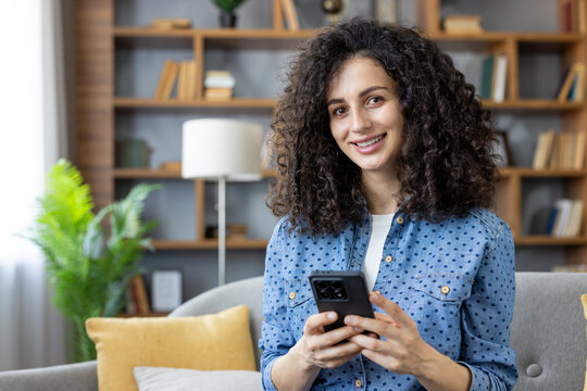 Young woman with curly hair sitting on a sofa, happily browsing her smartphone and looking at the camera, enjoying digital communication and connectivity in a cozy home environment - Powered by Adobe