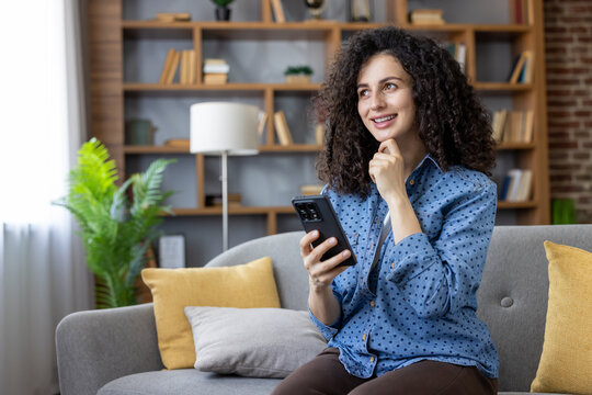 Woman with curly hair sitting on a comfortable couch, holding a smartphone, looking up and smiling while contemplating or planning, enjoying leisure time at home