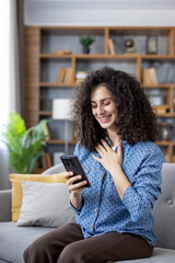 Young woman with curly hair sitting on a couch smiling and hand on chest as she reads a joyful...