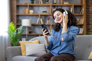 Woman with curly hair listening to music through headphones, holding smartphone, smiling and relaxing on a comfortable sofa in her living room at home