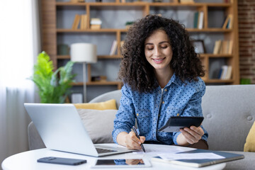 Young woman with curly hair calculating finances using a calculator and laptop, smiling while managing her budget at home, representing remote work and financial planning