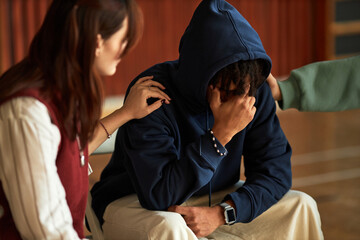 Black teenager boy sitting with head in hand being comforted by Caucasian teenager girl and another teenager, students supporting peer during emotional moment in psychology context