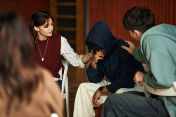 Multiethnic group of young adults comforting distressed Black man wearing hoodie, sitting in support circle during psychology class, students showing empathy and support through physical touch