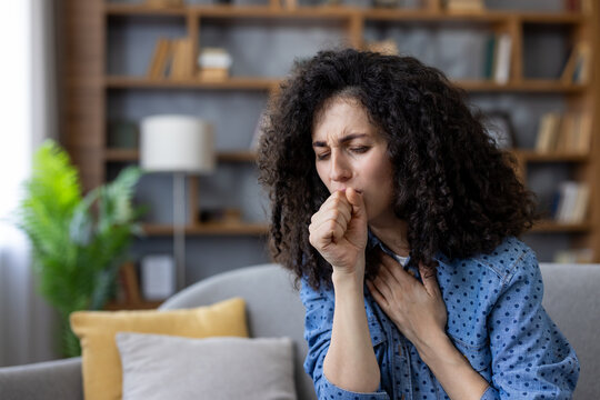 Woman with curly hair suffering from a painful cough, feeling unwell due to a cold, flu, allergy, or other respiratory illness while resting on a sofa in her home