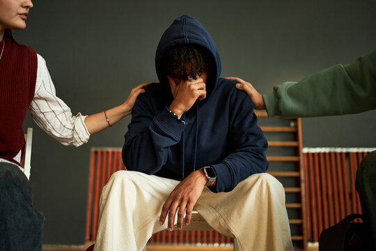 Black teenager boy sitting with head in hand showing distress, two students placing hands on his shoulders offering support, psychology group session setting, partial bodies visible