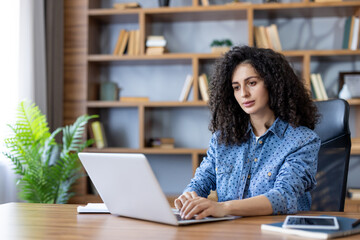 Young woman with curly hair focusing on her laptop while working remotely from a home office, surrounded by a bookshelf and desk with a plant and mobile phone