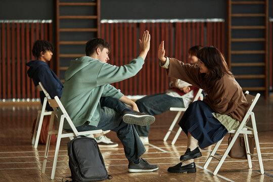 Group of multiethnic teenagers sitting on chairs in classroom setting, two teenagers giving high five while others observing, psychology students participating in interactive group activity