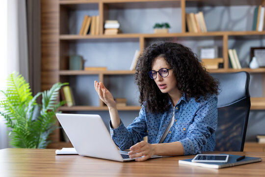 Young woman sitting at a desk, looking frustrated and confused at her laptop screen, encountering a technical problem or system error during a remote work session at her home office - Powered by Adobe