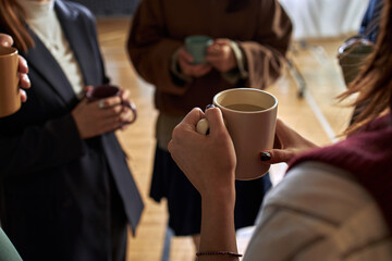Group of young adults and teenagers standing in circle holding mugs, engaging in conversation during psychology class, partial view focusing on hands and cups, indoor setting