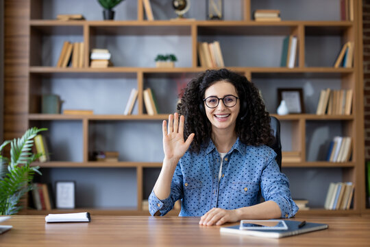 Woman in headset smiling and waving during a friendly video call from her home office, engaging in remote work with laptop on desk and bookshelf in the background