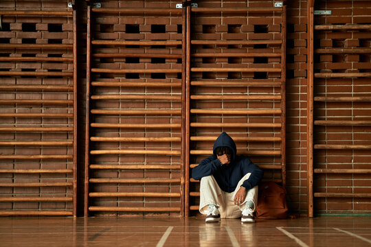 Teenage Black boy sitting alone on gym floor leaning against wooden wall bars wearing hooded sweatshirt resting head on hand appearing stressed