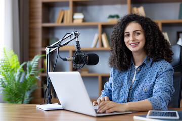 Young woman with curly hair hosting a podcast in her modern home office, smiling while using a laptop and professional microphone for remote work and online communication