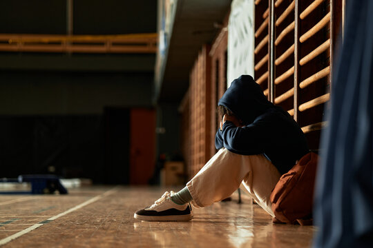 Teenager sitting alone on gym floor covering face with hands showing signs of emotional distress mental health struggle concept psychology students education environment