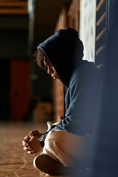 Black teenager sitting on floor with head down clasping hands appearing thoughtful in educational setting, context of mental health awareness