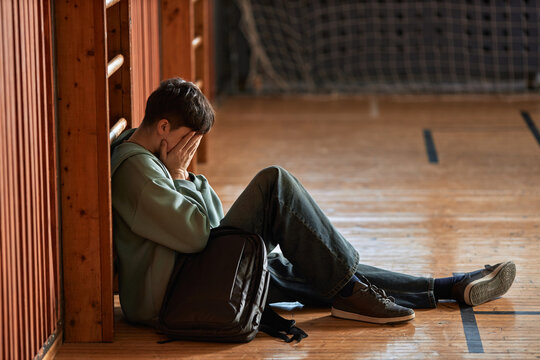 Teenage Caucasian boy sitting on gym floor leaning against wall covering face with hands showing stress or sadness with backpack beside him representing student mental health struggle