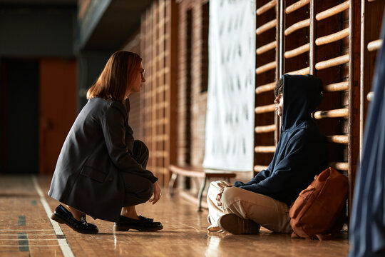 Caucasian young adult woman crouching and talking to seated teenager boy with curly hair and hoodie in gymnasium setting, both engaging in conversation near wooden wall bars