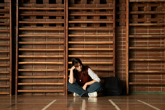 Caucasian teenage girl sitting on gym floor leaning against wooden wall bars holding head with hand looking down backpack beside her representing student experiencing stress