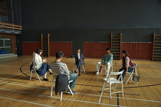 Group of multiethnic young adults and adult woman sitting in circle on chairs in sports hall discussing psychology topic during student workshop or group therapy session