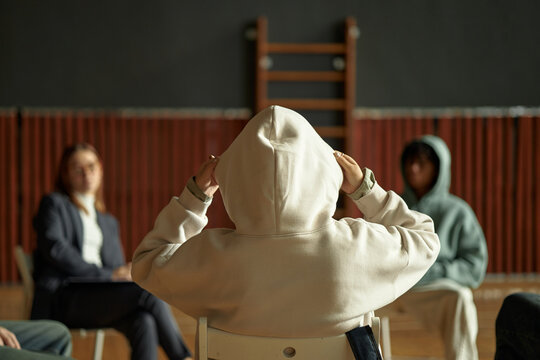 Teenager wearing hooded sweatshirt sitting with back to camera participating in group psychology session with Caucasian woman and Black teenager in background