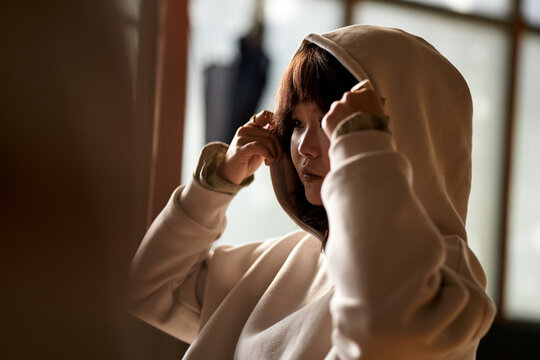 Asian female teenager adjusting hood while sitting indoors, focused expression visible, soft natural light illuminating face, student preparing for therapy session