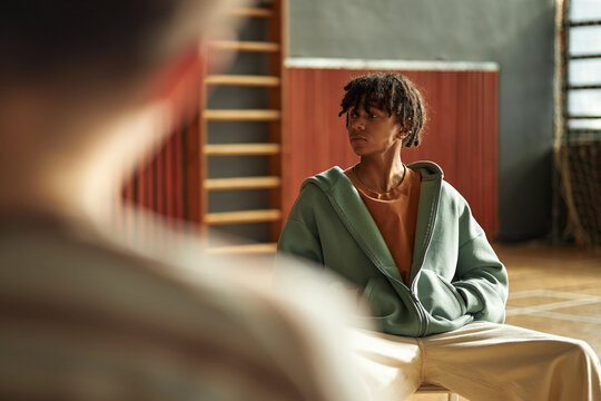 Black teenage boy sitting in classroom turning head to side listening to classmate during psychology lesson, wearing casual clothes, wooden gym equipment and window in background