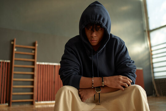Portrait of teenage Black boy sitting in gymnasium wearing hooded sweatshirt, hands clasped together, feeling stressed, therapy session concept 