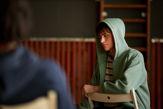 Teenage Caucasian boy sitting sideways on chair wearing hooded sweatshirt, looking away from camera, attending psychology class with another student visible in foreground