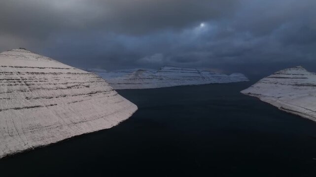 Aerial view of snow capped mountains rising from the dark ocean under a cloudy sky, creating a dramatic, stark contrast, Streymoy, Streymoy, Faroe Islands.