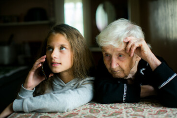 A girl speaks on the phone while an elderly woman watches her quietly, both sharing a calm moment in soft indoor light.