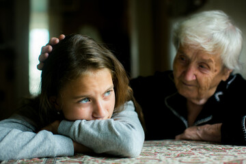 A tearful girl rests her head on her arms while an elderly woman gently comforts her, sharing a...