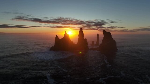 Aerial view of stark black sea stacks rise from the turbulent ocean, illuminated by the golden glow of the setting sun, Reynisfjara, Myrdalshreppur, Iceland.