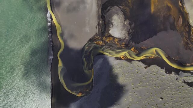 Aerial view of a vibrant yellow river snaking through dark volcanic sands, contrasting sharply with the green ocean waters, Grindavikurbaer, Iceland.
