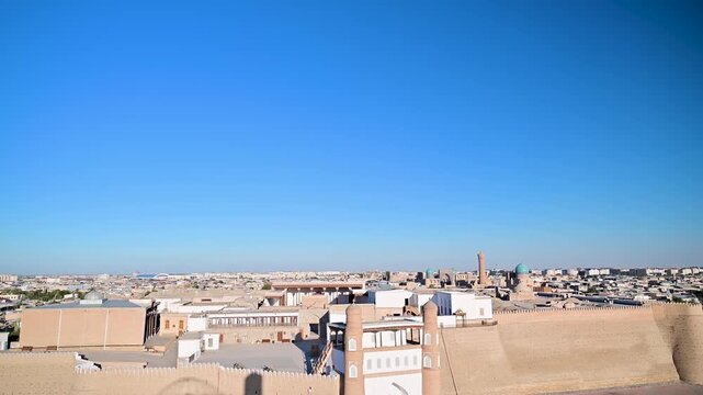 A high-angle aerial view of the Ark of Bukhara in Uzbekistan, showing the curved outer walls, entrance area, and surrounding historic center under warm evening light. 