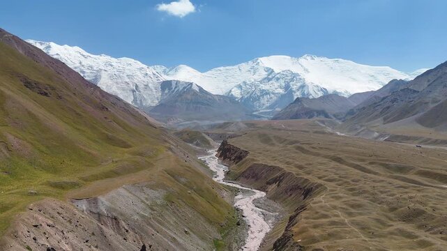 Drone footage captures a muddy river flowing through a dramatic mountain gorge, originating from the Alay Range. In the background, snow-covered peaks rise above the rugged terrain under clear skies.