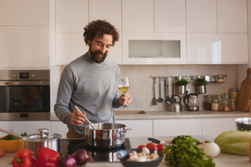 A man is actively cooking in his stylish kitchen, stirring a pot on the stove while holding a glass of wine. Fresh vegetables and herbs surround him, highlighting his focus on healthy eating.
