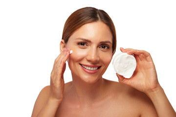 A woman with smooth skin is joyfully applying cream to her face while holding a jar of moisturizer. The background is bright and uncluttered, highlighting her beauty routine.