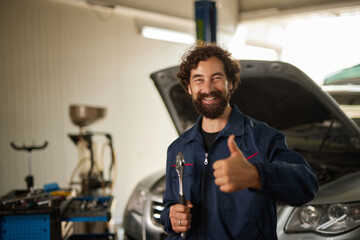 A mechanic stands in a well-lit auto repair shop smiling and giving a thumbs up. The vehicle's hood is open, showing the ongoing repair work. Tools are visible nearby.
