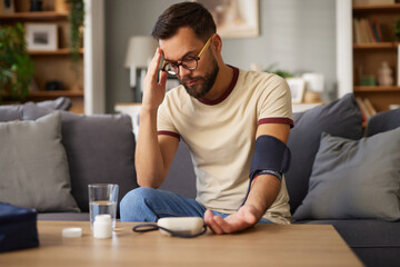 A man sits on a couch in a cozy living room, using a blood pressure monitor while looking worried....
