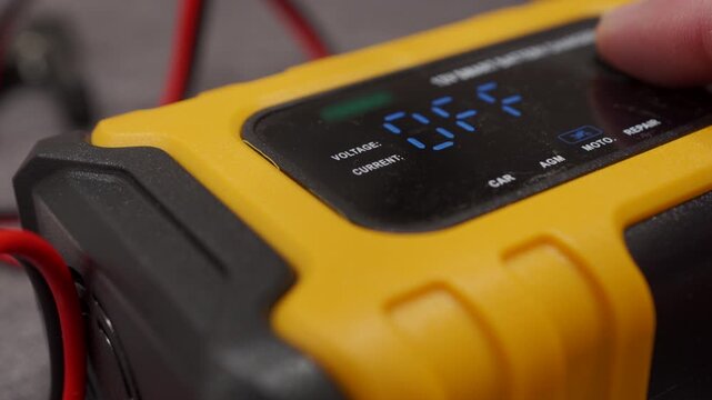 A yellow and black plastic car battery charger is operated. A finger presses the mode selection button on the digital display, which makes a short, dry click.