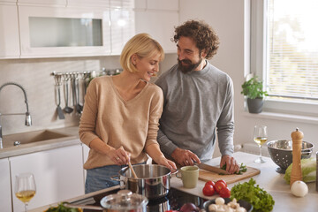 A man and woman are happily cooking in a modern kitchen. They are chopping vegetables, stirring a pot, and sharing smiles amidst fresh ingredients and sunlight.