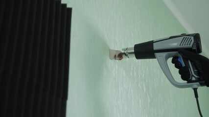 Close-up of repairman heating and carefully removing old adhesive tape residue from wall using professional hot air blower tool, preparing surface for new soundproofing panels. Shooting in slow motion