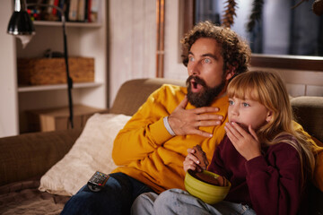 A father and daughter sit closely on a couch, both surprised and engaged as they watch a suspenseful movie in their cozy living room during evening hours.