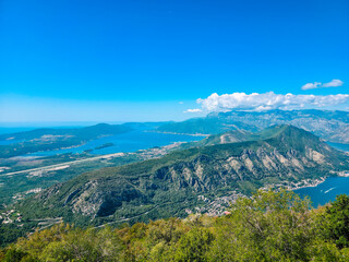 Scenic panoramic view of the Bay of Kotor in Montenegro, showing deep blue water, high green mountains, winding coastline and distant islands under a bright clear sky. A peaceful Mediterranean summer 