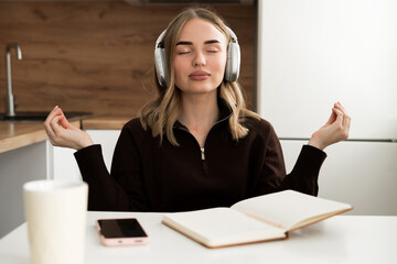 Young Woman in headphones meditating, doing yoga breathing in the kitchen at home. Calm serene student taking break.