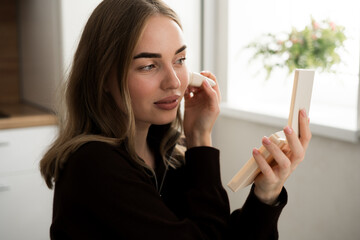 Cute young woman applying blush on face with brush looking in mirror sitting near desk at home