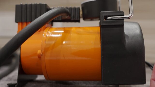 An orange and black metal and plastic car air compressor is running. The motor produces a loud, low-pitched, rhythmic chugging and vibrating sound as it pumps air.